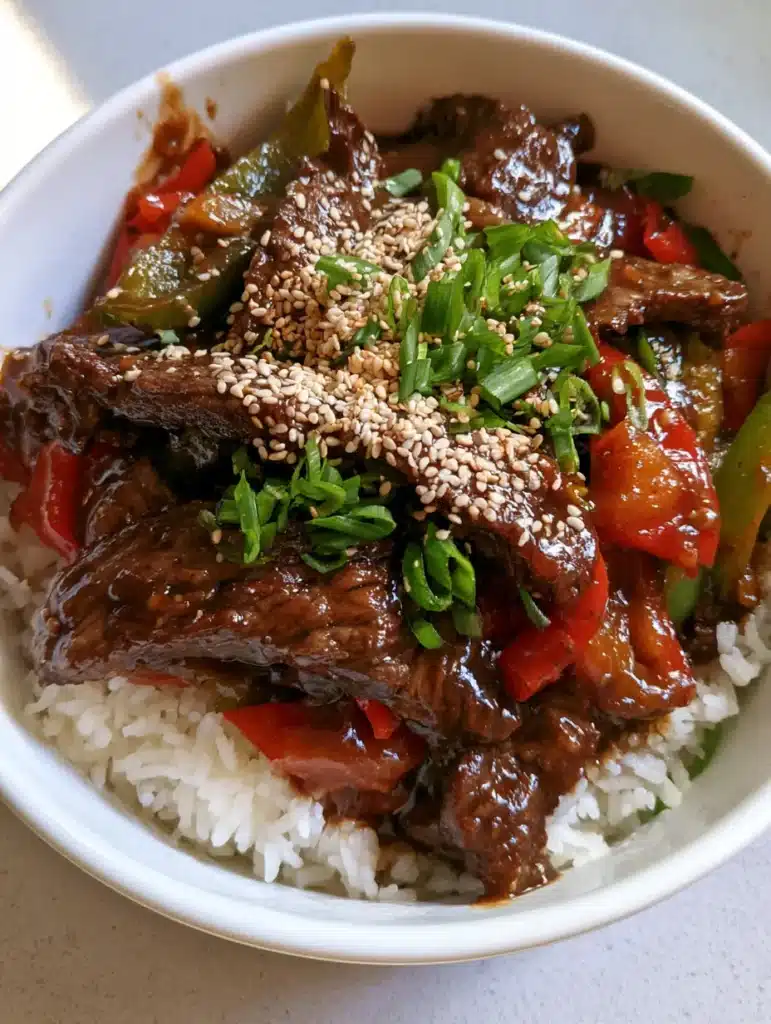 A close up view of Slow Cooker Pepper Steak showing the seared beef strips and glossy dark brown sauce garnished with sesame seeds.