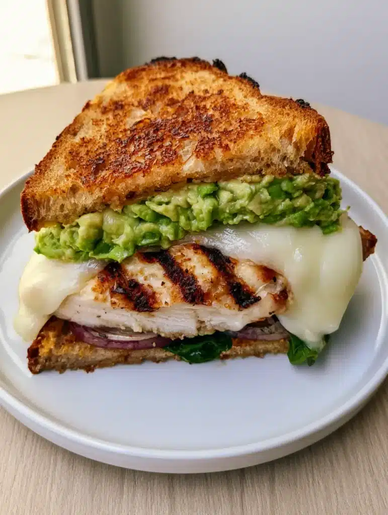 Chicken Avocado Melt with Toasted Sourdough and Char-Grilled Chicken 1 A Chicken Avocado Melt on sourdough bread sliced in half on a white plate with a wood background.