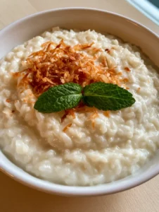 A white ceramic bowl filled with thick and velvety coconut rice pudding, topped with golden-brown toasted coconut flakes and fresh mint leaves. (Coconut Rice Pudding)